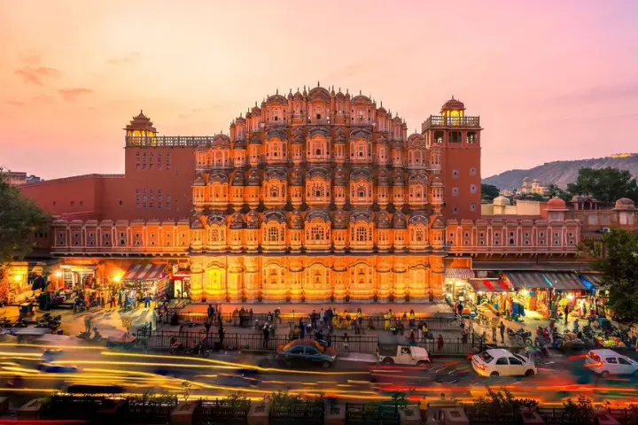 Hawa Mahal in Jaipur, India, at sunset. The palace, made of red and pink sandstone, is lit up against an orange and pink sky. Below, the street is bustling with people and cars, their headlights and taillights creating streaks of light due to a long exposure.